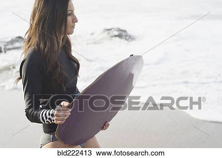 Stock Image - Caucasian woman holding skimboard at beach. Fotosearch