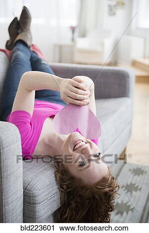 Stock Image - Caucasian woman laying on sofa holding valentine heart. Fotosearch