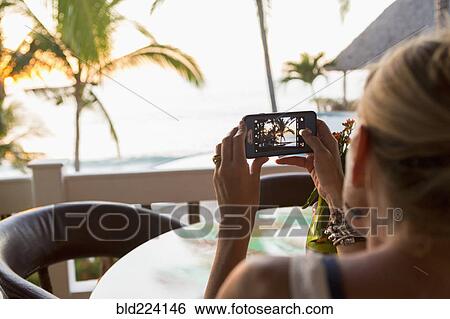 Stock Photograph - Caucasian woman photographing sunset at beach. Fotosearch