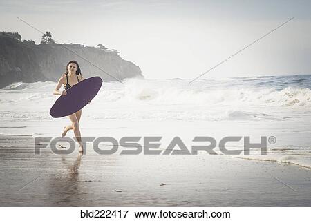 Stock Photo - Caucasian woman running with skimboard at beach. Fotosearch