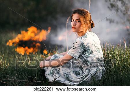 Stock Image - Caucasian woman sitting near fire in rural field. Fotosearch