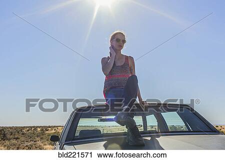Stock Image - Caucasian woman sitting on roof of car in desert. Fotosearch