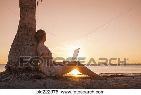 Stock Photograph - Caucasian woman with laptop leaning on palm tree at sunset. Fotosearch