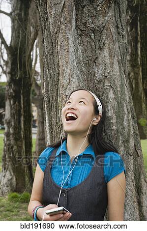 Stock Image - Chinese girl listening to headphones against tree. Fotosearch