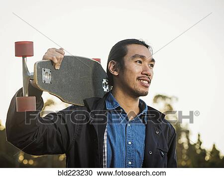 Stock Image - Chinese man carrying skateboard outdoors. Fotosearch