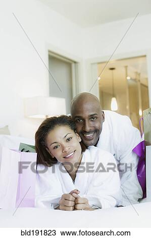 Stock Image - Couple in bathrobes laying on bed with shopping bags. Fotosearch