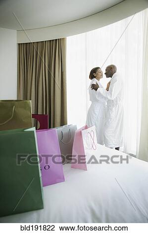 Stock Image - Couple kissing in bathrobes with shopping bags on bed. Fotosearch