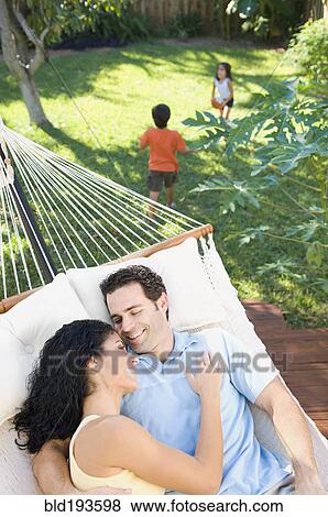 Stock Photo - Couple relaxing in hammock with children playing in background. Fotosearch