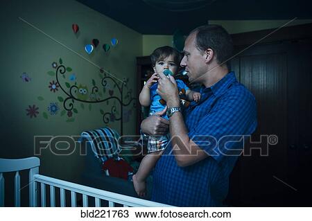 Stock Image - Father giving baby son pacifier in nursery. Fotosearch