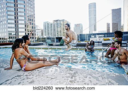 Stock Image - Friends watching man do cannonball dive into urban swimming pool. Fotosearch