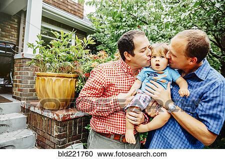 Stock Image - Gay fathers kissing baby son outdoors. Fotosearch