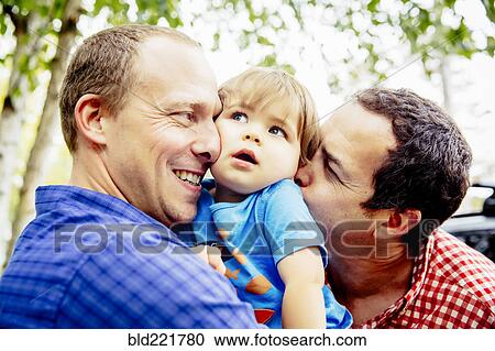 Stock Image - Gay fathers kissing baby son outdoors. Fotosearch