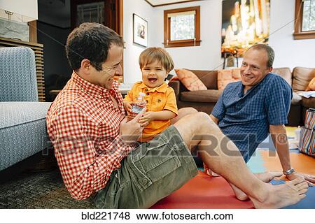 Stock Photo - Gay fathers playing with baby son in living room. Fotosearch
