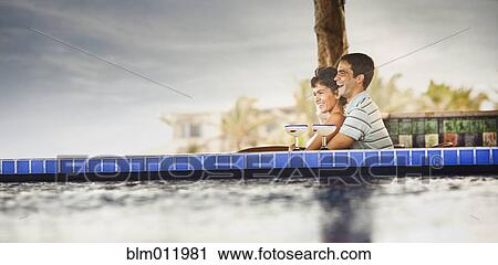 Stock Image - Hispanic couple drinking cocktails at poolside. Fotosearch