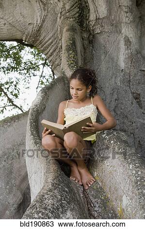 Stock Image - Hispanic girl reading a book and sitting on large tree roots. Fotosearch