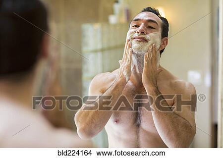 Picture - Hispanic man applying shaving cream to face in mirror. Fotosearch