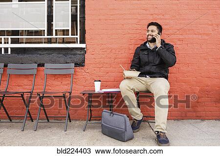 Stock Image - Hispanic man eating bowl of food on sidewalk using cell phone. Fotosearch