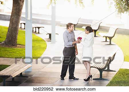 Stock Image - Hispanic man giving girlfriend flowers in park. Fotosearch