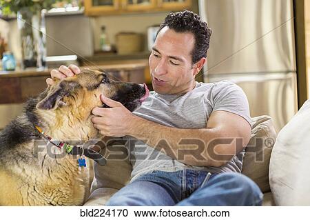 Stock Image - Hispanic man laying on sofa petting dog. Fotosearch