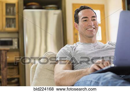 Stock Photo - Hispanic man laying on sofa using laptop. Fotosearch