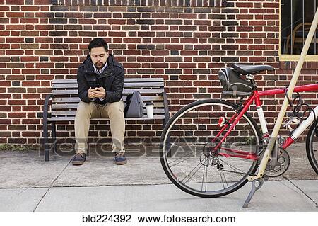 Hispanic man sitting on city bench using cell phone View Large Photo Image Stock Image - Hispanic man sitting on city bench using cell phone. Fotosearch