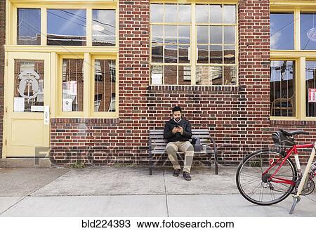 Stock Image - Hispanic man sitting on city bench using cell phone. Fotosearch