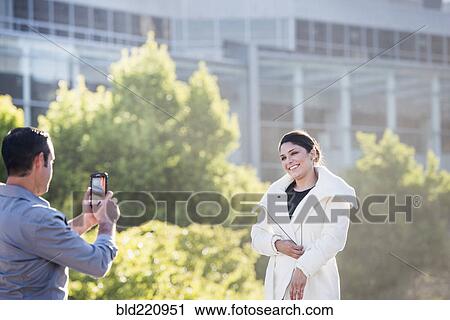 Stock Image - Hispanic man taking cell phone picture of girlfriend in city. Fotosearch