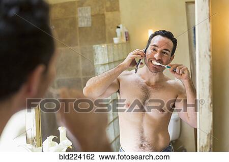 Stock Image - Hispanic man using cell phone and brushing teeth in mirror. Fotosearch