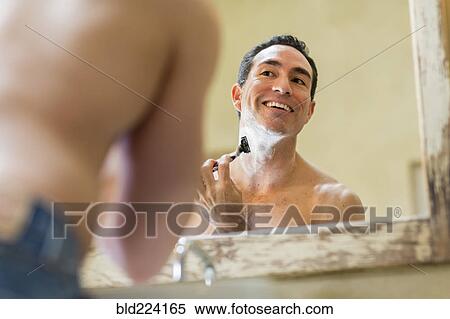 Stock Photography - Hispanic man using razor to shave face in mirror. Fotosearch