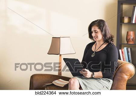 Picture - Hispanic woman sitting on armchair reading digital tablet. Fotosearch