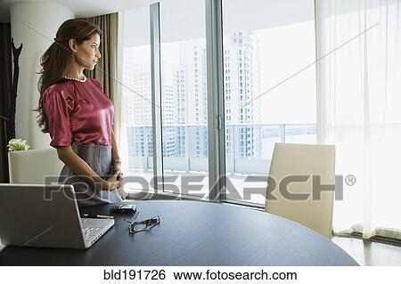 Stock Photograph - Hispanic woman standing next to laptop and looking out window. Fotosearch