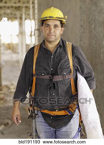 Stock Image - Hispanic worker holding blueprints on construction site. Fotosearch