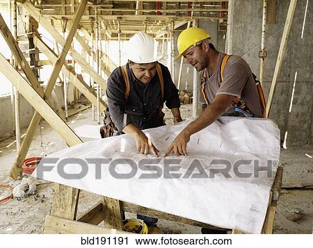 Stock Image - Hispanic workers looking at blueprints on construction site. Fotosearch