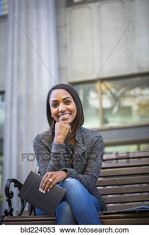 Stock Image - Indian woman sitting on city bench holding booklet. Fotosearch