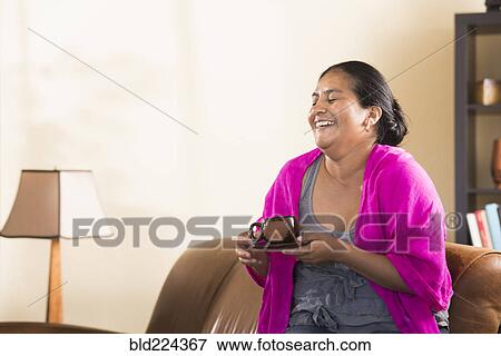 Laughing Hispanic woman drinking coffee in livingroom View Large Photo Image Stock Photo - Laughing Hispanic woman drinking coffee in livingroom. Fotosearch