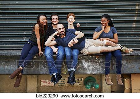 Stock Image - Man laying in laps of friends on loading dock. Fotosearch