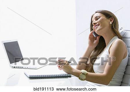 Stock Image - Mixed race businesswoman talking on cell phone at desk. Fotosearch
