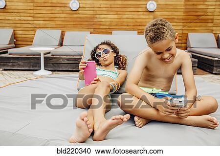 Stock Image - Mixed race children taking selfie with cell phones on patio. Fotosearch
