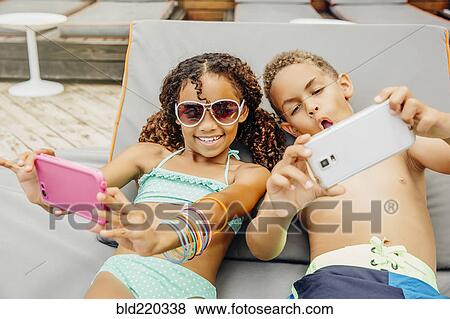 Stock Photo - Mixed race children taking selfie with cell phones on patio. Fotosearch