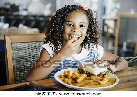 Stock Image - Mixed race girl eating sandwich at table. Fotosearch