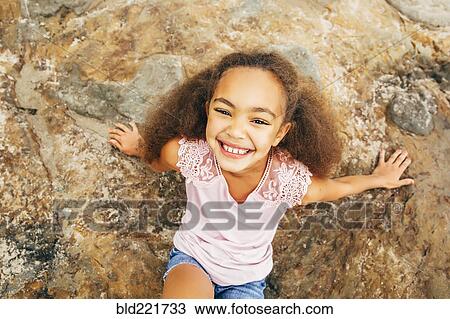 Stock Image - Mixed race girl smiling on rock outdoors. Fotosearch