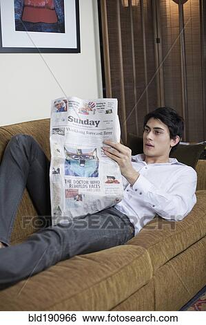 Stock Photograph - Mixed race man laying on sofa reading newspaper. Fotosearch