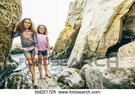 Stock Photo - Mixed race sisters smiling on rocks. Fotosearch