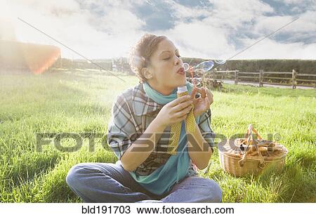 Stock Image - Mixed race woman blowing bubbles in sunny field. Fotosearch