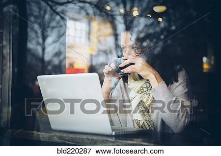 Stock Photo - Mixed race woman drinking coffee and using laptop in cafe. Fotosearch