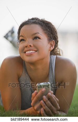 Picture - Mixed race woman laying in grass holding cell phone. Fotosearch