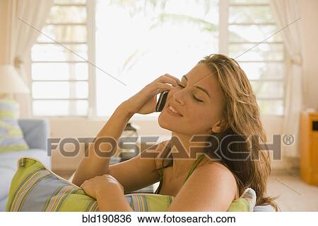 Stock Photograph - Mixed race woman sitting on sofa in living room talking on cell phone. Fotosearch