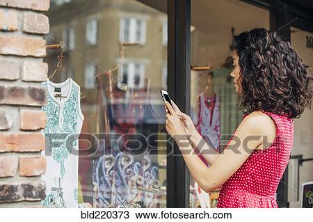 Stock Image - Mixed race woman taking cell phone photograph of clothes in store window. Fotosearch