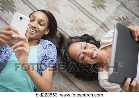 Stock Image - Mother and daughter laying on carpet using cell phone and digital tablet. Fotosearch