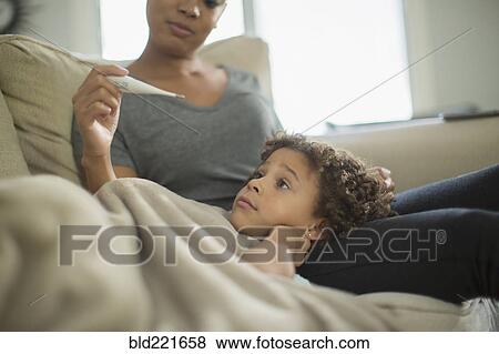 Stock Photo - Mother measuring temperature of daughter on sofa. Fotosearch